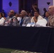 A formal conference or summit is taking place in a large ballroom or event hall. Several attendees are seated at a front-row table with a black tablecloth, including a military officer in tan uniform and civilians wearing blue lanyards. A photographer is visible in the background capturing the event.