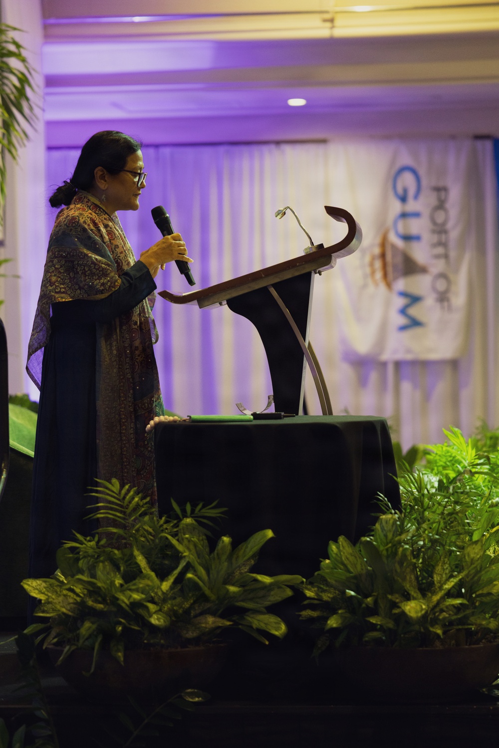 A woman is speaking at a formal event, standing at a podium while holding a handheld microphone. She is dressed in a dark patterned outfit with a shawl and glasses. A banner in the background partially reads 'Port of Guam,' indicating this is an official or governmental event held in Guam.