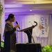 A woman is speaking at a formal event, standing at a podium while holding a handheld microphone. She is dressed in a dark patterned outfit with a shawl and glasses. A banner in the background partially reads 'Port of Guam,' indicating this is an official or governmental event held in Guam.