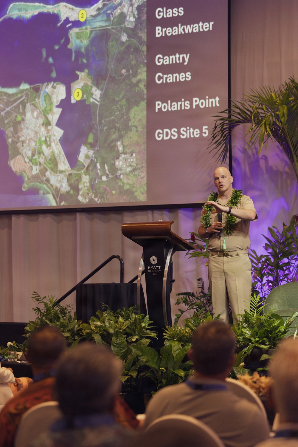 A bald male speaker in tan/khaki military-style uniform and a green lei is presenting at a podium in a Hyatt Regency conference venue. Behind him is a projected slide showing a satellite aerial map with numbered points and text listing 'Glass Breakwater,' 'Gantry Cranes,' 'Polaris Point,' and 'GDS Site 5.' An audience is seated in the foreground watching the presentation.