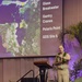 A bald male speaker in tan/khaki military-style uniform and a green lei is presenting at a podium in a Hyatt Regency conference venue. Behind him is a projected slide showing a satellite aerial map with numbered points and text listing 'Glass Breakwater,' 'Gantry Cranes,' 'Polaris Point,' and 'GDS Site 5.' An audience is seated in the foreground watching the presentation.