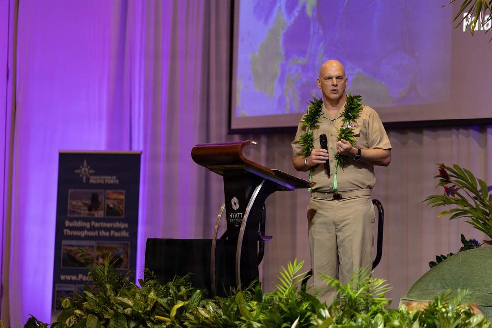 A bald military officer in tan/khaki uniform is delivering a speech at a podium during what appears to be a Pacific ports conference. He is wearing a traditional Hawaiian lei and holding a microphone, standing at a Hyatt Regency venue. A projection screen displaying a map is visible behind him.