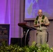 A bald military officer in tan/khaki uniform is delivering a speech at a podium during what appears to be a Pacific ports conference. He is wearing a traditional Hawaiian lei and holding a microphone, standing at a Hyatt Regency venue. A projection screen displaying a map is visible behind him.