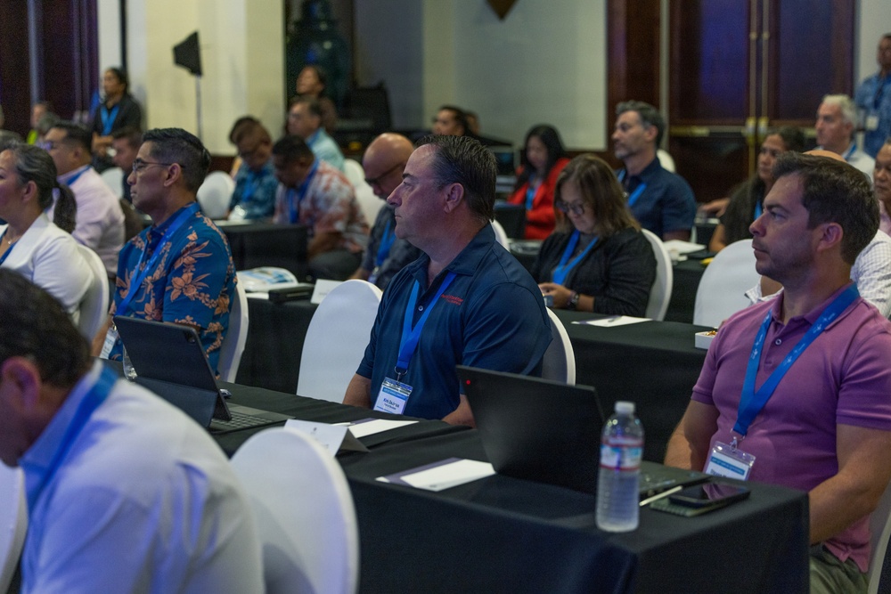 A professional conference or seminar is taking place in a formal meeting room, with approximately 20-30 attendees seated at black-draped tables. Participants are wearing name badge lanyards in blue and appear to be attentively listening to a presentation. The setting suggests a structured business or organizational event, likely in a tropical or Pacific region based on attendee attire.