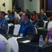 A professional conference or seminar is taking place in a formal meeting room, with approximately 20-30 attendees seated at black-draped tables. Participants are wearing name badge lanyards in blue and appear to be attentively listening to a presentation. The setting suggests a structured business or organizational event, likely in a tropical or Pacific region based on attendee attire.