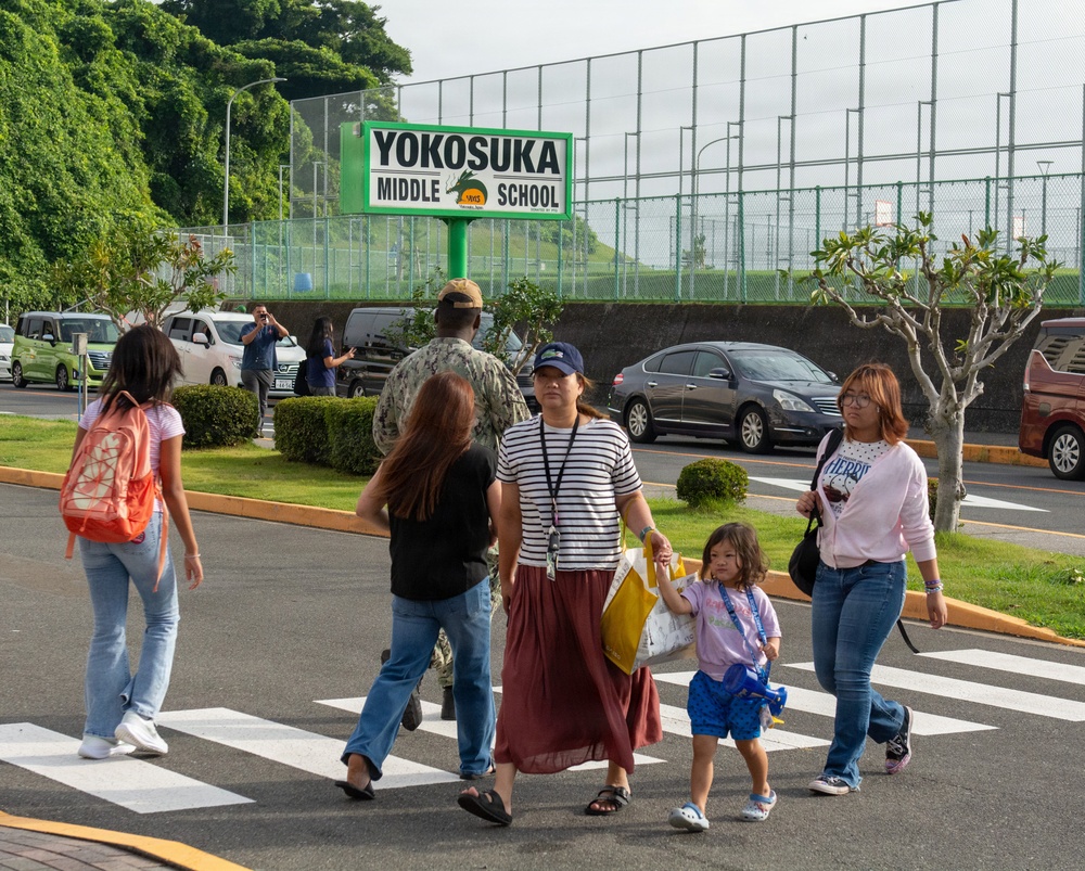 A group of people are crossing a zebra crosswalk in front of Yokosuka Middle School, a Department of Defense Dependents School located in Yokosuka, Japan. The scene appears to be during a school arrival or dismissal period, with adults and children walking together. A green school sign prominently displays 'Yokosuka Middle School' in the background.