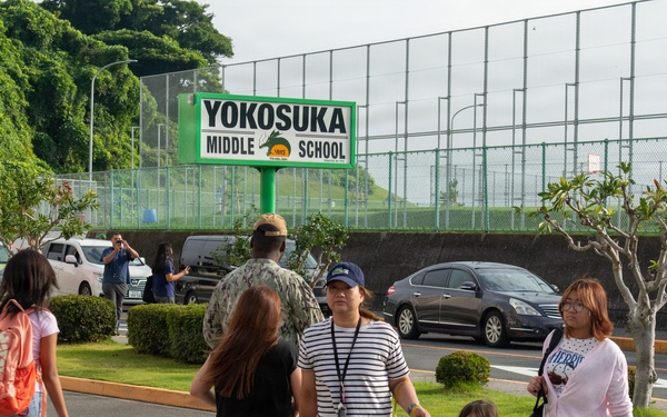 A group of people are crossing a zebra crosswalk in front of Yokosuka Middle School, a Department of Defense Dependents School located in Yokosuka, Japan. The scene appears to be during a school arrival or dismissal period, with adults and children walking together. A green school sign prominently displays 'Yokosuka Middle School' in the background.