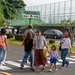 A group of people are crossing a zebra crosswalk in front of Yokosuka Middle School, a Department of Defense Dependents School located in Yokosuka, Japan. The scene appears to be during a school arrival or dismissal period, with adults and children walking together. A green school sign prominently displays 'Yokosuka Middle School' in the background.