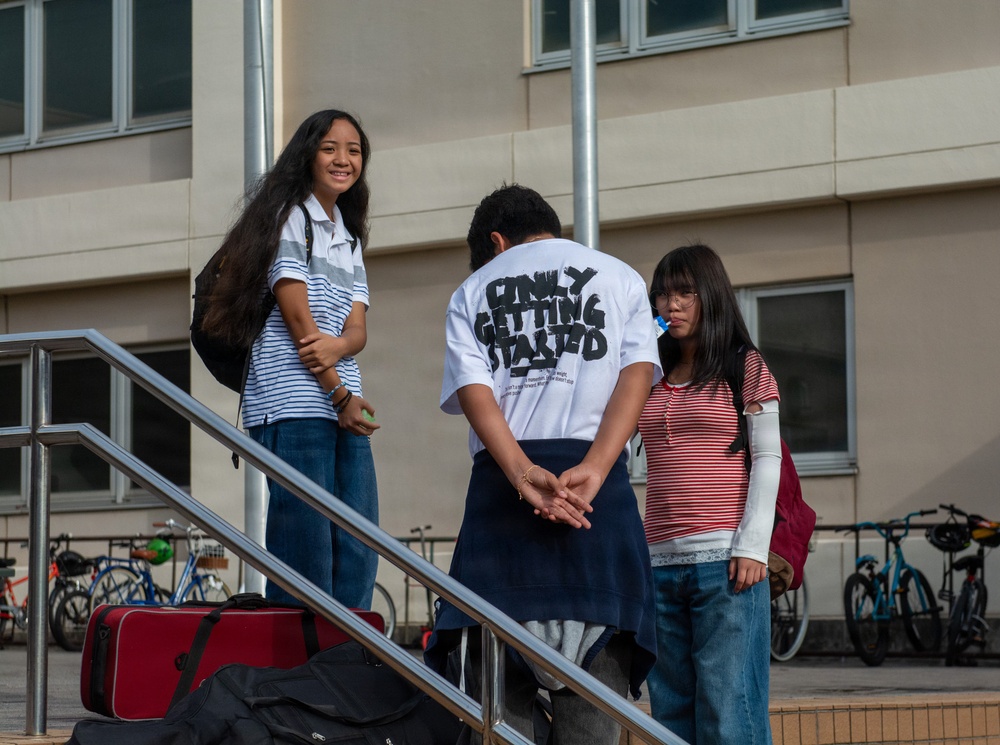 Three young people are gathered on an outdoor staircase in front of a modern building, appearing to be in conversation. One person with their back to the camera wears a white t-shirt reading 'ONLY GETTING STARTED.' The setting appears to be a school or university campus, with bicycles parked in the background and luggage on the steps.