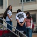 Three young people are gathered on an outdoor staircase in front of a modern building, appearing to be in conversation. One person with their back to the camera wears a white t-shirt reading 'ONLY GETTING STARTED.' The setting appears to be a school or university campus, with bicycles parked in the background and luggage on the steps.