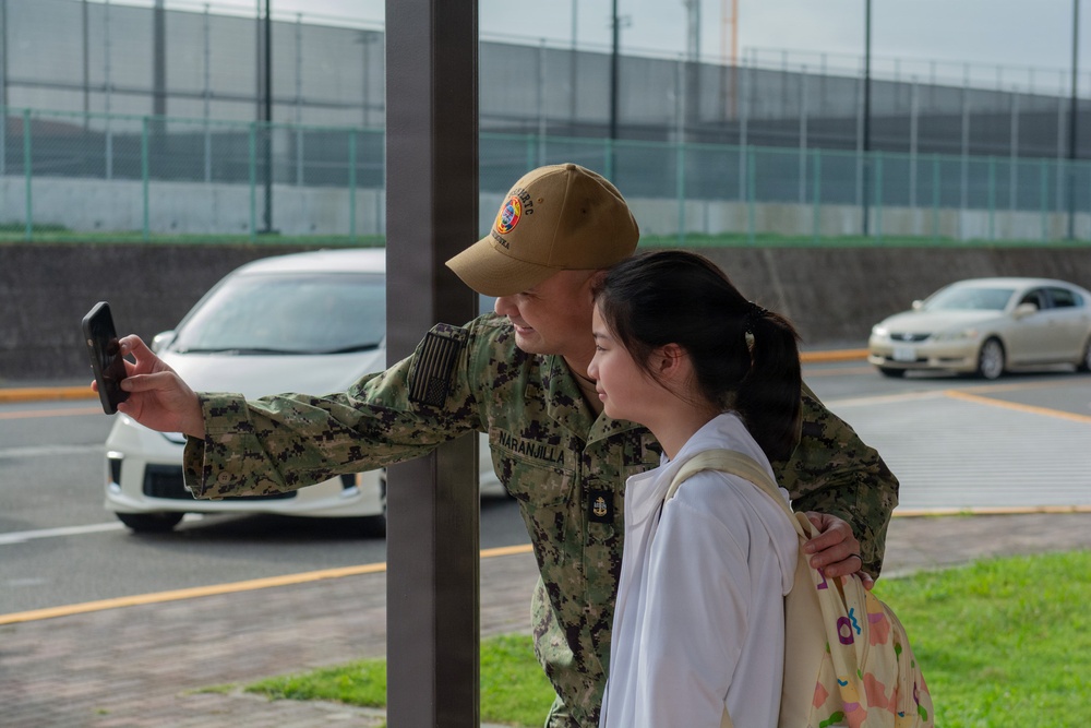 A U.S. Navy service member in digital camouflage uniform is taking a selfie with a young woman near a parking area. The sailor, whose name tape reads 'NARANJILLA,' has their arm around the woman while extending a smartphone outward. The setting appears to be a military installation, likely in Japan based on the vehicle styles and infrastructure.