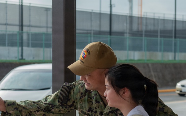 A U.S. Navy service member in digital camouflage uniform is taking a selfie with a young woman near a parking area. The sailor, whose name tape reads 'NARANJILLA,' has their arm around the woman while extending a smartphone outward. The setting appears to be a military installation, likely in Japan based on the vehicle styles and infrastructure.