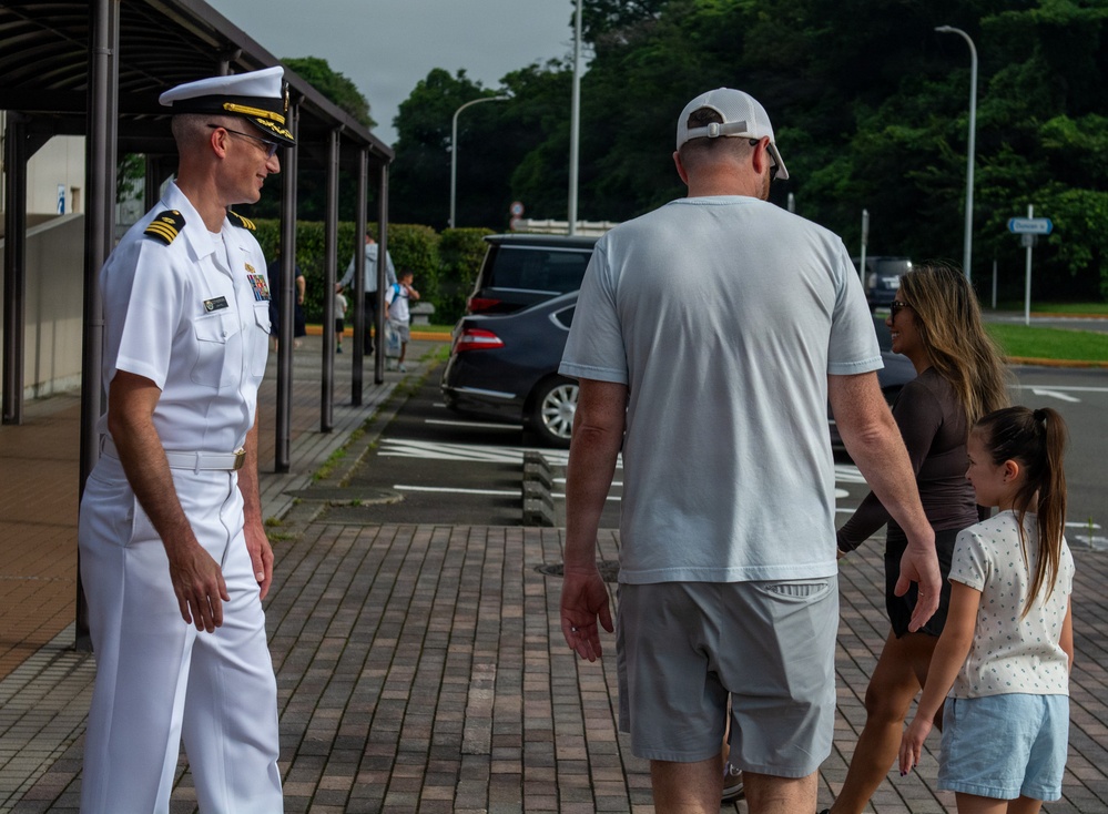 A U.S. Navy officer in white dress uniform stands on a brick walkway, appearing to greet or interact with a civilian family consisting of a tall adult male, a woman, and a young girl. The setting appears to be a military installation, likely in Japan based on the architectural style and landscaping. The officer displays captain or commander rank insignia with gold shoulder boards.