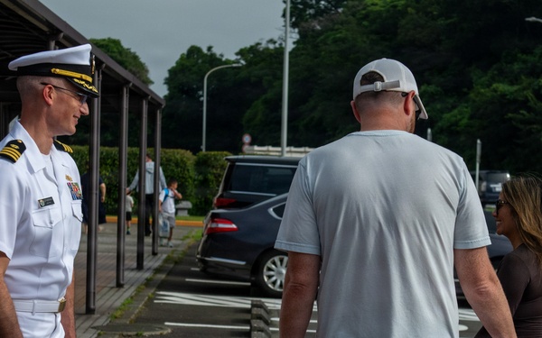 A U.S. Navy officer in white dress uniform stands on a brick walkway, appearing to greet or interact with a civilian family consisting of a tall adult male, a woman, and a young girl. The setting appears to be a military installation, likely in Japan based on the architectural style and landscaping. The officer displays captain or commander rank insignia with gold shoulder boards.