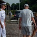 A U.S. Navy officer in white dress uniform stands on a brick walkway, appearing to greet or interact with a civilian family consisting of a tall adult male, a woman, and a young girl. The setting appears to be a military installation, likely in Japan based on the architectural style and landscaping. The officer displays captain or commander rank insignia with gold shoulder boards.