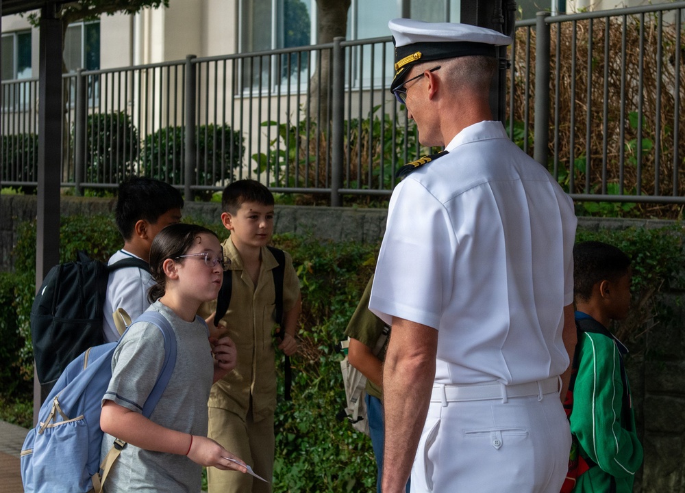 A naval officer in white dress uniform is engaged in conversation with a group of school-aged children on a sidewalk outside a building. The officer, wearing a white cover (cap) with gold insignia, appears to be interacting with students who are carrying backpacks. The setting suggests a school visit or community outreach event.