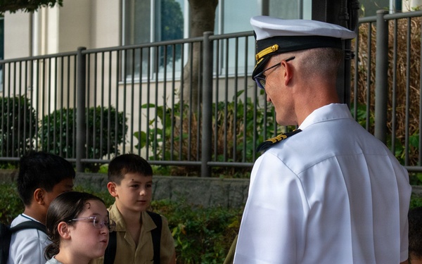 A naval officer in white dress uniform is engaged in conversation with a group of school-aged children on a sidewalk outside a building. The officer, wearing a white cover (cap) with gold insignia, appears to be interacting with students who are carrying backpacks. The setting suggests a school visit or community outreach event.