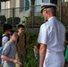 A naval officer in white dress uniform is engaged in conversation with a group of school-aged children on a sidewalk outside a building. The officer, wearing a white cover (cap) with gold insignia, appears to be interacting with students who are carrying backpacks. The setting suggests a school visit or community outreach event.