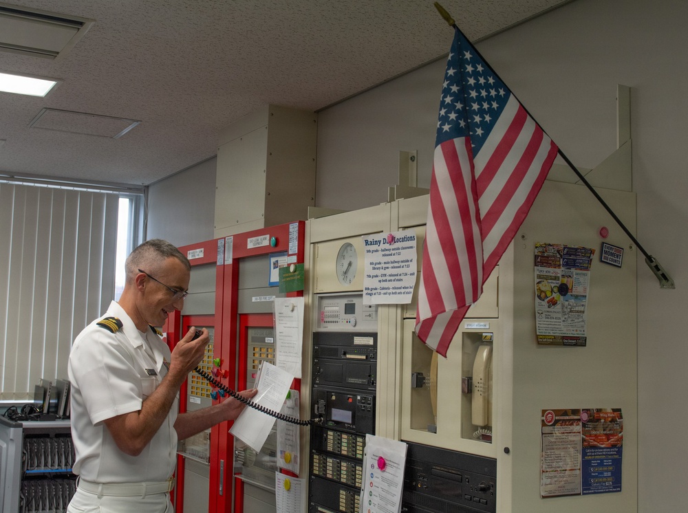 A uniformed military officer in white dress uniform is using a handheld radio/intercom while holding papers in what appears to be a school or institutional administrative office. An American flag is mounted on the wall behind him, and the room contains fire alarm panels, communication equipment, and various posted notices. The setting appears to be a front office or security/communications hub of a school facility.