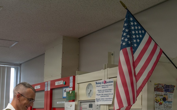 A uniformed military officer in white dress uniform is using a handheld radio/intercom while holding papers in what appears to be a school or institutional administrative office. An American flag is mounted on the wall behind him, and the room contains fire alarm panels, communication equipment, and various posted notices. The setting appears to be a front office or security/communications hub of a school facility.