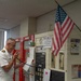 A uniformed military officer in white dress uniform is using a handheld radio/intercom while holding papers in what appears to be a school or institutional administrative office. An American flag is mounted on the wall behind him, and the room contains fire alarm panels, communication equipment, and various posted notices. The setting appears to be a front office or security/communications hub of a school facility.
