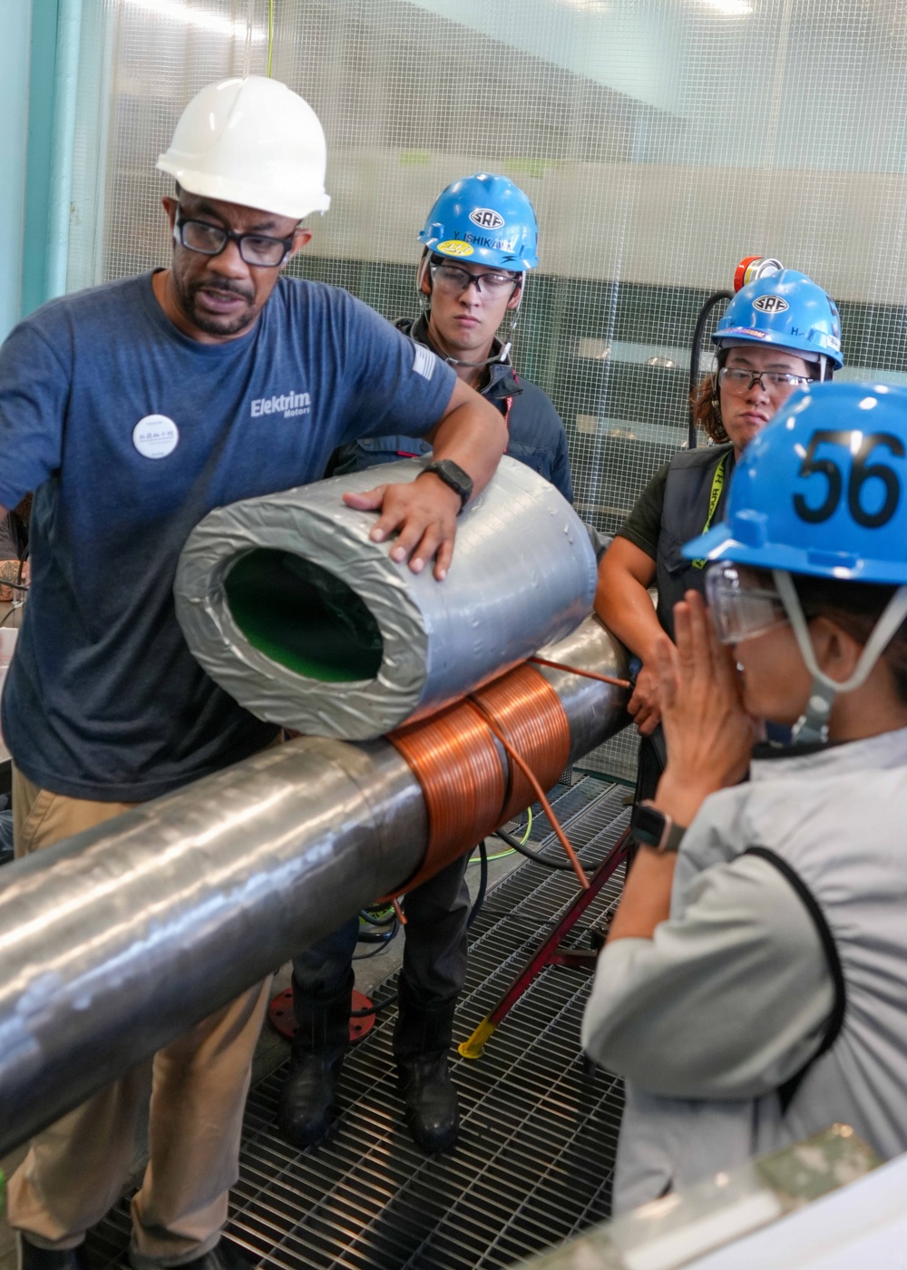 A group of workers in an industrial facility are examining a cylindrical component wrapped in metallic tape with a copper coil winding around it. The person in the foreground wearing a white hard hat and an 'Elektrim Motors' shirt appears to be demonstrating or explaining the component to colleagues wearing blue 'SRF' branded hard hats. The setting appears to be a technical or manufacturing environment with grated metal flooring.