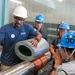 A group of workers in an industrial facility are examining a cylindrical component wrapped in metallic tape with a copper coil winding around it. The person in the foreground wearing a white hard hat and an 'Elektrim Motors' shirt appears to be demonstrating or explaining the component to colleagues wearing blue 'SRF' branded hard hats. The setting appears to be a technical or manufacturing environment with grated metal flooring.