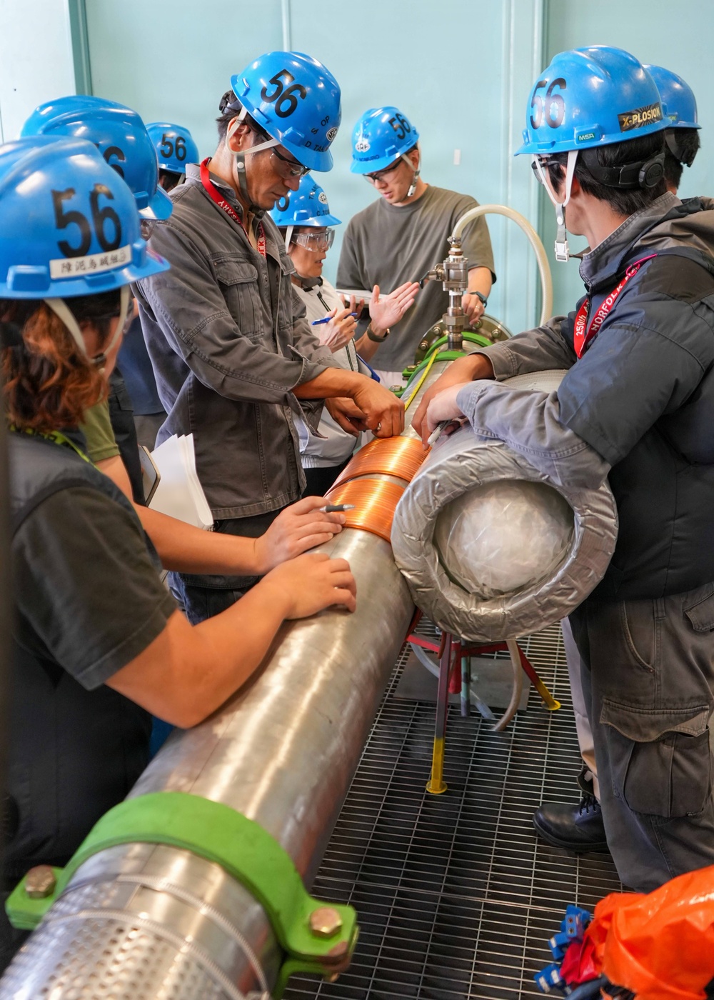 A group of workers or trainees wearing blue hard hats numbered '56' are gathered around a large industrial pipe with copper coil winding, appearing to conduct a hands-on inspection or training exercise. The setting appears to be an industrial facility or training center with grated flooring and teal-colored walls. An instructor or senior technician appears to be demonstrating or explaining something about the pipe assembly to the group.