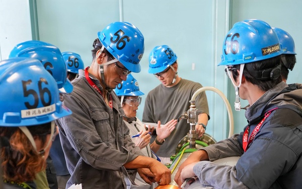 A group of workers or trainees wearing blue hard hats numbered '56' are gathered around a large industrial pipe with copper coil winding, appearing to conduct a hands-on inspection or training exercise. The setting appears to be an industrial facility or training center with grated flooring and teal-colored walls. An instructor or senior technician appears to be demonstrating or explaining something about the pipe assembly to the group.