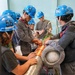 A group of workers or trainees wearing blue hard hats numbered '56' are gathered around a large industrial pipe with copper coil winding, appearing to conduct a hands-on inspection or training exercise. The setting appears to be an industrial facility or training center with grated flooring and teal-colored walls. An instructor or senior technician appears to be demonstrating or explaining something about the pipe assembly to the group.