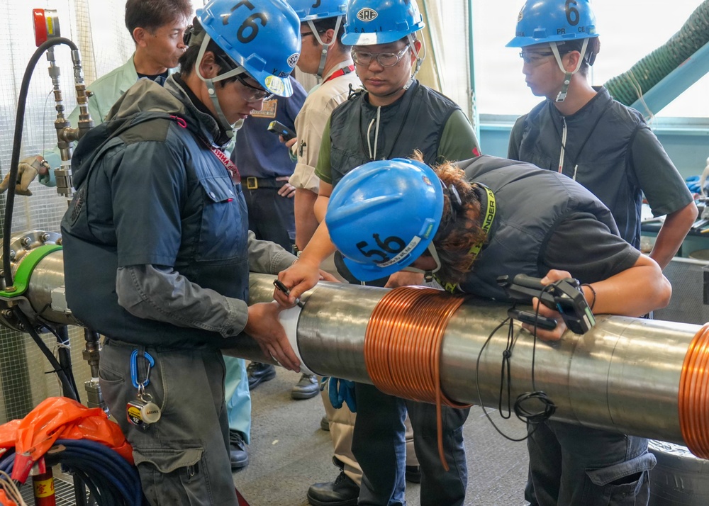 A group of workers wearing blue hard hats and safety glasses are gathered around a large metallic cylindrical pipe or shaft that has a copper coil wrapped around it. The team appears to be inspecting, measuring, or performing technical work on the equipment in what looks like an industrial or engineering facility. At least one person is leaning in closely to examine or mark the pipe while others observe.