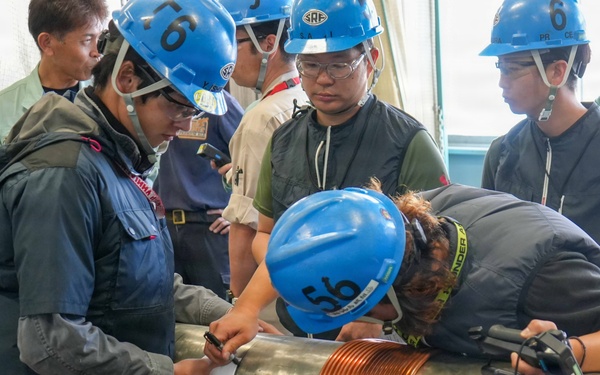 A group of workers wearing blue hard hats and safety glasses are gathered around a large metallic cylindrical pipe or shaft that has a copper coil wrapped around it. The team appears to be inspecting, measuring, or performing technical work on the equipment in what looks like an industrial or engineering facility. At least one person is leaning in closely to examine or mark the pipe while others observe.