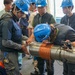 A group of workers wearing blue hard hats and safety glasses are gathered around a large metallic cylindrical pipe or shaft that has a copper coil wrapped around it. The team appears to be inspecting, measuring, or performing technical work on the equipment in what looks like an industrial or engineering facility. At least one person is leaning in closely to examine or mark the pipe while others observe.