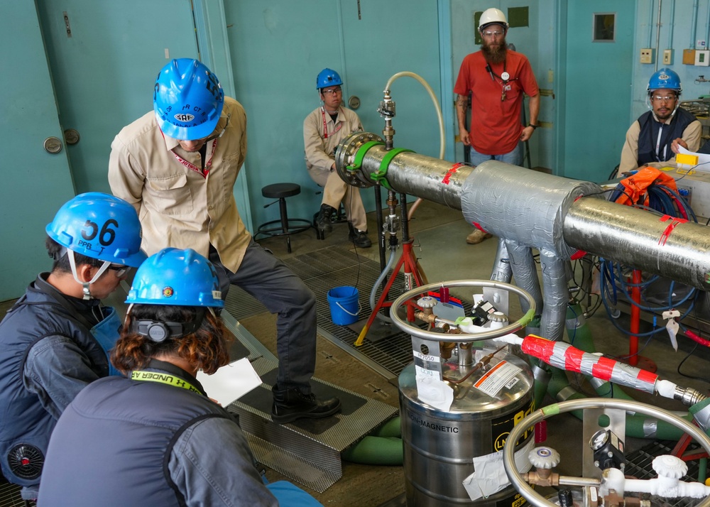 A group of approximately six workers in a technical or industrial facility are gathered around large insulated piping and specialized equipment. Several workers wear blue hard hats and safety glasses, with one wearing a white hard hat, suggesting a supervised technical operation or experiment. The setting appears to be a laboratory or test facility, with visible cryogenic or pressurized system components.