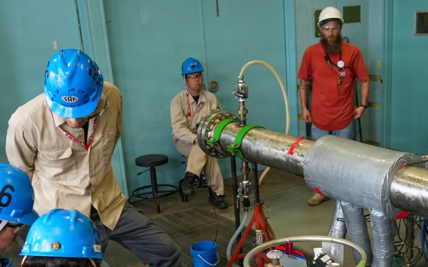 A group of approximately six workers in a technical or industrial facility are gathered around large insulated piping and specialized equipment. Several workers wear blue hard hats and safety glasses, with one wearing a white hard hat, suggesting a supervised technical operation or experiment. The setting appears to be a laboratory or test facility, with visible cryogenic or pressurized system components.