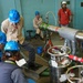 A group of approximately six workers in a technical or industrial facility are gathered around large insulated piping and specialized equipment. Several workers wear blue hard hats and safety glasses, with one wearing a white hard hat, suggesting a supervised technical operation or experiment. The setting appears to be a laboratory or test facility, with visible cryogenic or pressurized system components.