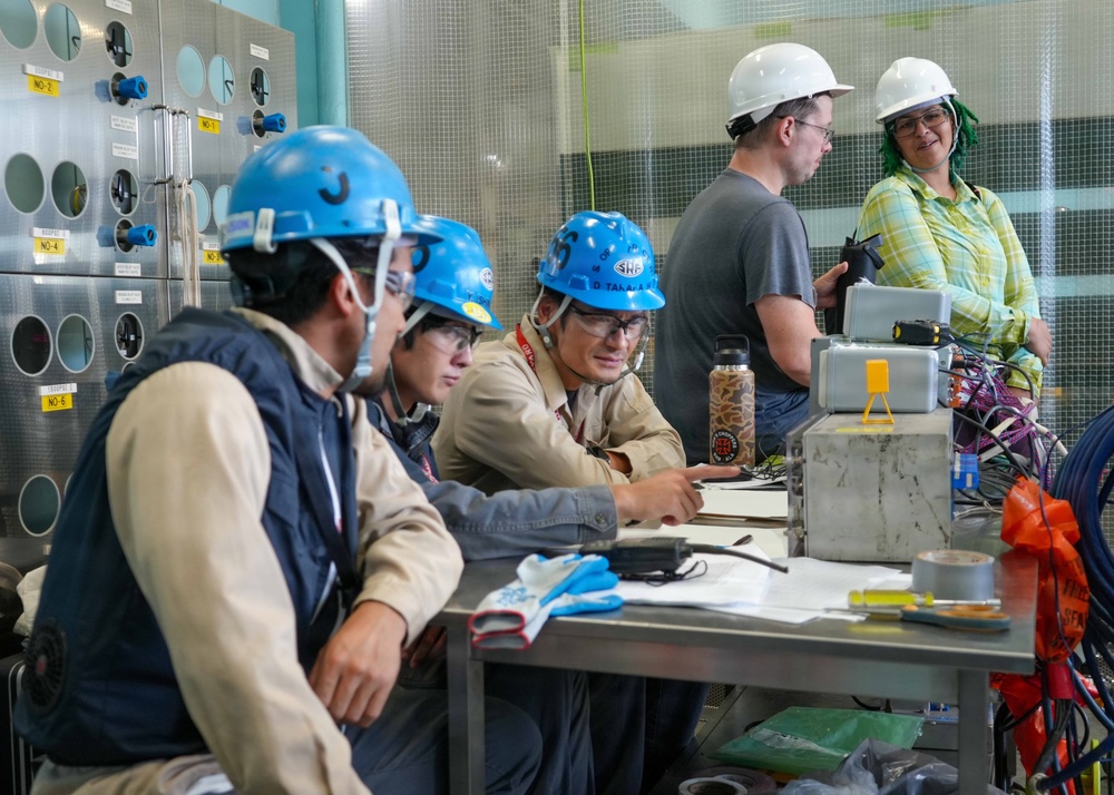 A group of workers in hard hats and safety gear are gathered around a metal worktable in what appears to be an industrial or technical facility. Three workers wearing blue hard hats are reviewing documents and discussing work, while two others wearing white hard hats stand nearby. The setting includes a panel wall with numbered ports or connectors and various tools and equipment on the table.