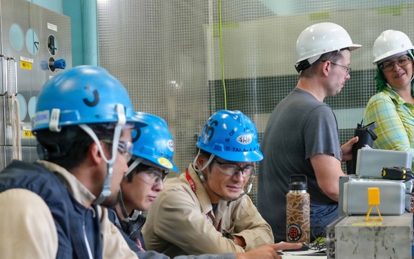 A group of workers in hard hats and safety gear are gathered around a metal worktable in what appears to be an industrial or technical facility. Three workers wearing blue hard hats are reviewing documents and discussing work, while two others wearing white hard hats stand nearby. The setting includes a panel wall with numbered ports or connectors and various tools and equipment on the table.
