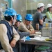 A group of workers in hard hats and safety gear are gathered around a metal worktable in what appears to be an industrial or technical facility. Three workers wearing blue hard hats are reviewing documents and discussing work, while two others wearing white hard hats stand nearby. The setting includes a panel wall with numbered ports or connectors and various tools and equipment on the table.