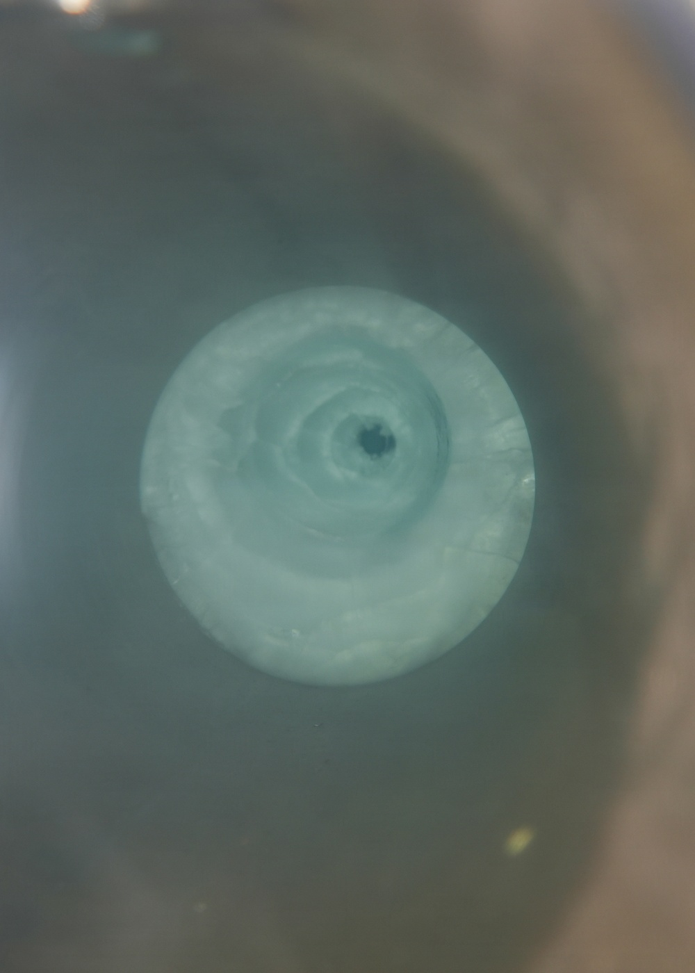 The image shows a top-down view of a swirling vortex or whirlpool in a liquid-filled container. The liquid appears to be a pale blue-green color, with a distinct spiral pattern converging toward a dark central drain point. The surrounding container appears to be a basin or tub with a brownish rim visible on the right edge.