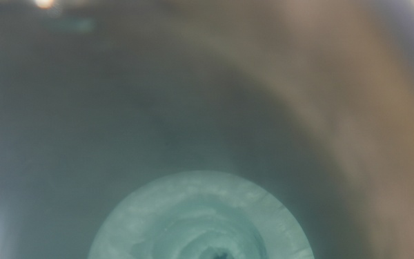 The image shows a top-down view of a swirling vortex or whirlpool in a liquid-filled container. The liquid appears to be a pale blue-green color, with a distinct spiral pattern converging toward a dark central drain point. The surrounding container appears to be a basin or tub with a brownish rim visible on the right edge.