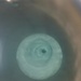 The image shows a top-down view of a swirling vortex or whirlpool in a liquid-filled container. The liquid appears to be a pale blue-green color, with a distinct spiral pattern converging toward a dark central drain point. The surrounding container appears to be a basin or tub with a brownish rim visible on the right edge.