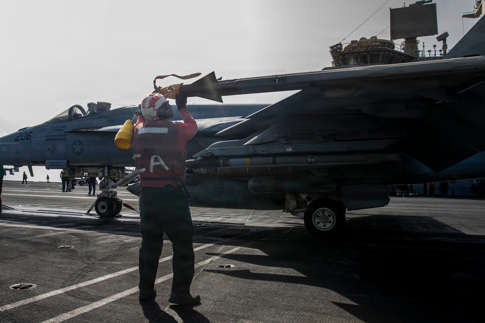 A U.S. Navy flight deck crew member in a red jersey is performing a pre-flight or post-flight inspection on a carrier-based jet aircraft, likely an F/A-18 Hornet or Super Hornet, on the flight deck of an aircraft carrier. The crew member appears to be checking or removing a control surface cover or streamer near the aircraft's tail section. Additional aircraft and deck personnel are visible in the background.
