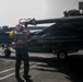 A U.S. Navy flight deck crew member in a red jersey is performing a pre-flight or post-flight inspection on a carrier-based jet aircraft, likely an F/A-18 Hornet or Super Hornet, on the flight deck of an aircraft carrier. The crew member appears to be checking or removing a control surface cover or streamer near the aircraft's tail section. Additional aircraft and deck personnel are visible in the background.