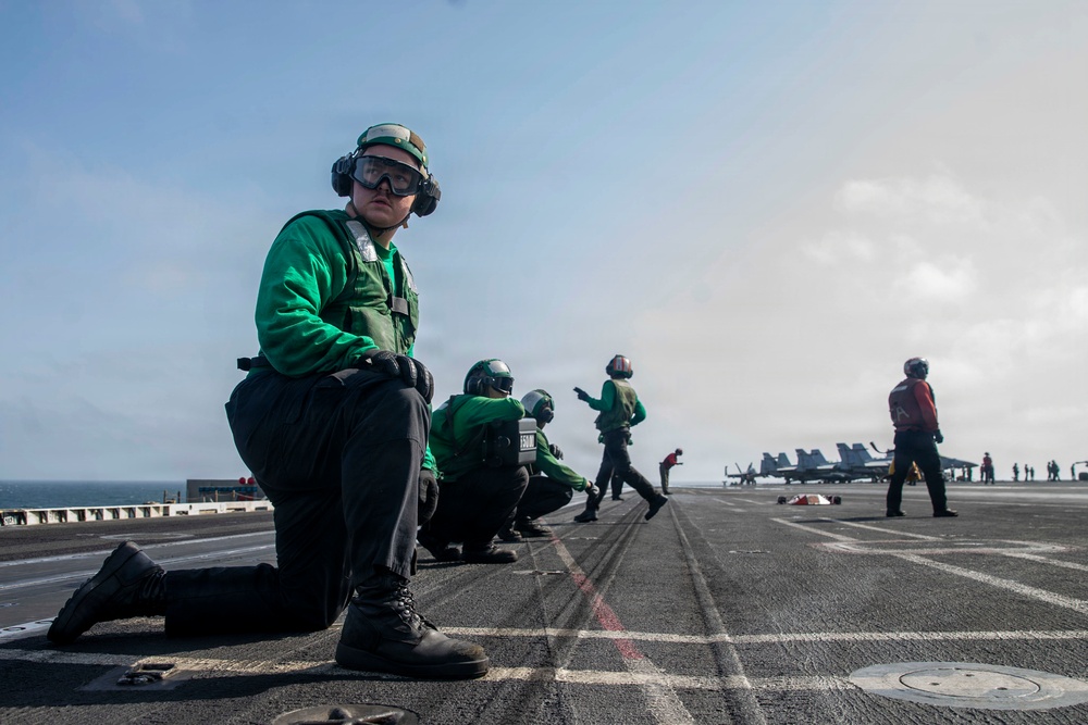 The image shows flight deck crew members aboard a U.S. Navy aircraft carrier, wearing green jerseys and protective gear including goggles and hearing protection. Personnel are positioned in various stances along the flight deck, with military jet aircraft visible in the background. The scene depicts standard carrier flight deck operations with crew in their designated roles.