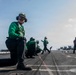 The image shows flight deck crew members aboard a U.S. Navy aircraft carrier, wearing green jerseys and protective gear including goggles and hearing protection. Personnel are positioned in various stances along the flight deck, with military jet aircraft visible in the background. The scene depicts standard carrier flight deck operations with crew in their designated roles.