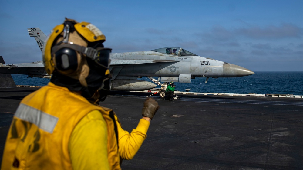 A U.S. Navy aircraft carrier flight deck scene showing an F/A-18 Hornet or Super Hornet marked '201' from squadron VFA-14 positioned on the deck, apparently preparing for launch. A yellow-vested flight deck crew member (shooter or handler) is visible in the foreground giving hand signals, while a green-vested crew member crouches near the aircraft's nose gear. The ocean horizon is visible in the background under a partly cloudy sky.