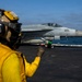 A U.S. Navy aircraft carrier flight deck scene showing an F/A-18 Hornet or Super Hornet marked '201' from squadron VFA-14 positioned on the deck, apparently preparing for launch. A yellow-vested flight deck crew member (shooter or handler) is visible in the foreground giving hand signals, while a green-vested crew member crouches near the aircraft's nose gear. The ocean horizon is visible in the background under a partly cloudy sky.