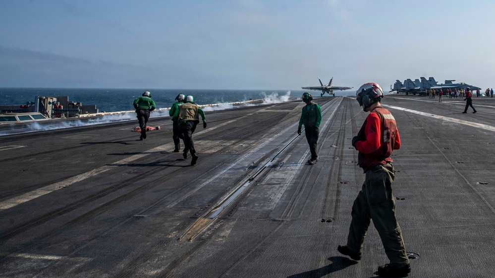 A carrier flight deck operation is underway, showing a jet aircraft — likely an F/A-18 Hornet or Super Hornet — launching via catapult from the bow of a naval aircraft carrier at sea. Multiple deck crew members in color-coded jerseys are visible performing their assigned roles. Additional parked aircraft and personnel are visible in the background on the starboard side of the deck.