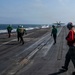 A carrier flight deck operation is underway, showing a jet aircraft — likely an F/A-18 Hornet or Super Hornet — launching via catapult from the bow of a naval aircraft carrier at sea. Multiple deck crew members in color-coded jerseys are visible performing their assigned roles. Additional parked aircraft and personnel are visible in the background on the starboard side of the deck.